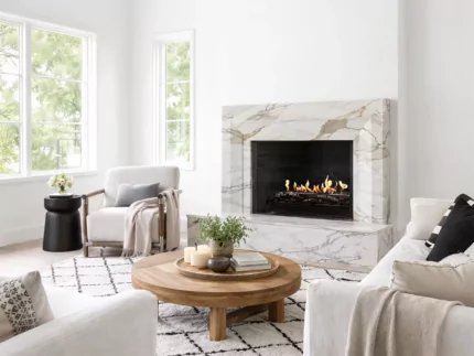 Bright, modern living room with white vaulted ceilings, a marble fireplace featuring an EcoSmart Fire Heritage Grate with a lit flame, neutral upholstered seating, a round wood coffee table, and a patterned rug, filled with natural light from tall windows.