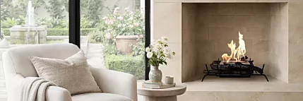 Cozy modern living room with a light upholstered armchair beside a lit stone fireplace, large black-framed windows, and a patterned area rug.